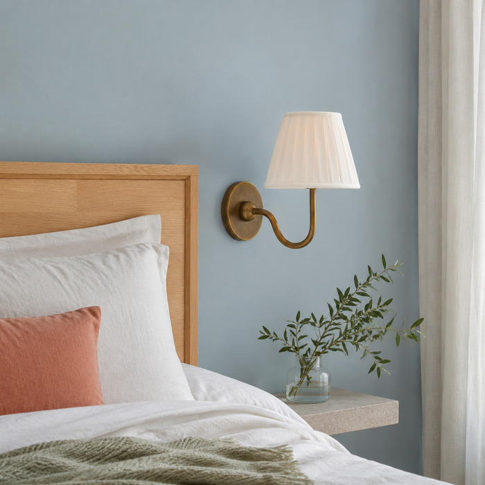 Een witte, geplooide Baylor-wandlamp naast natuurlijk linnen beddengoed in een rustige, blauwe slaapkamer.