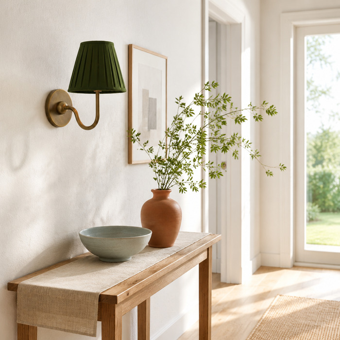 Groene, geplooide Baylor-wandlamp boven een licht eikenhouten console in een zonnige hal.